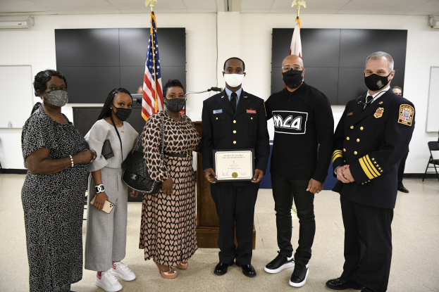 Eine Gruppe maskierter Menschen, die vor Fahnen stehen und eine Person in der Mitte ein Zertifikat hält, wahrscheinlich bei einer Abschlussfeier.