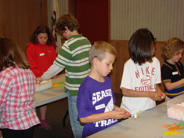 Kinder vor Tischen mit verschiedenen Gegenständen darauf, mit einer Flagge, Türen und einer Wand im Hintergrund.
