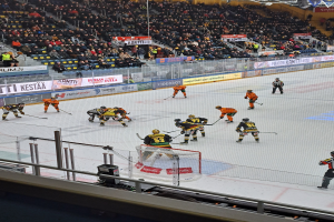 Eine Gruppe von Menschen, die Hockey auf einem Eisring mit einem Netz und einem umliegenden Zaun spielen, mit Zuschauern im Hintergrund und Bannern mit Text, unter hellen Stadionlichtern.