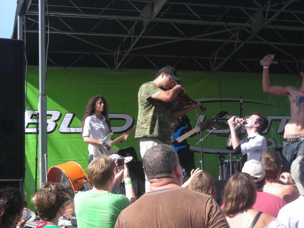 Eine Gruppe von Musikern performs auf der Bühne mit verschiedenen Instrumenten, während das Publikum vor einem Podium steht und die Musik genießt. Ein großer schwarzer Lautsprecher ist auf der linken Seite positioniert und ein grünes Banner ist im Hintergrund sichtbar.