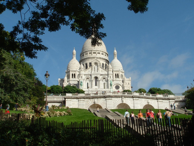 Ein weißes Gebäude, umgeben von einem Zaun, Gras, Bäumen und Menschen mit Stangen im Hintergrund.