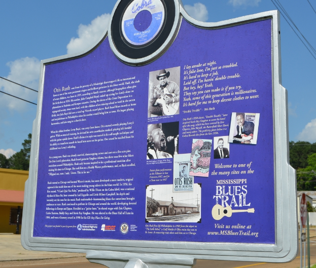 Eine Infotafel, Gebäude, ein Schuppen, Bäume, Stromkabel und ein bewölkter Himmel.