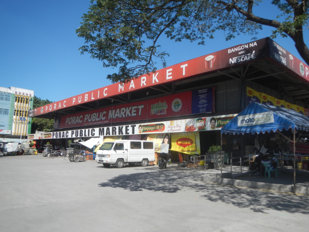 Öffentlicher Markt mit einem vor ihm geparkten Van, umgeben von Gebäuden, Geschäften, Schildern, Fahrzeugen auf der Straße, Menschen unter einem Zelt sitzend, Geländern, Bäumen und einem klaren blauen Himmel.