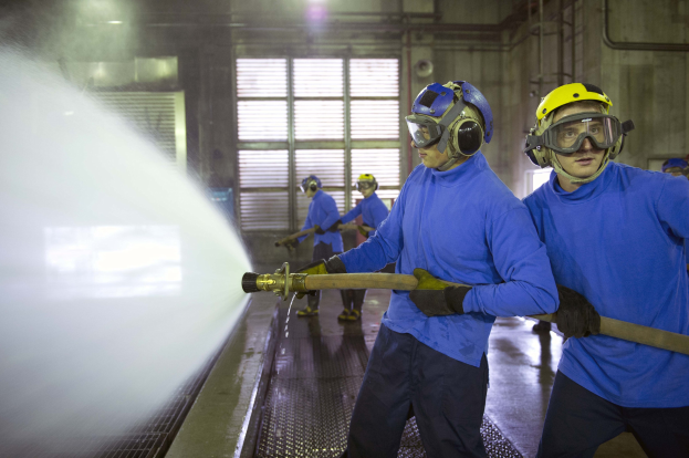 Gruppe von Männern in blauen Hemden und gelben Helmen, die an Maschinen arbeiten, wobei einer Wasser auf den Boden sprüht, in einer Fabrikumgebung mit sichtbaren Rohren, Fenstern und Lampen.