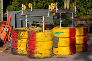 Ein Haufen gelber und roter Reifen auf einem Schmutzfeld umgeben von verstreuten Metallgegenständen, Brettern und anderem Schutt, mit Bäumen, Pflanzen und einer Hütte im Hintergrund.