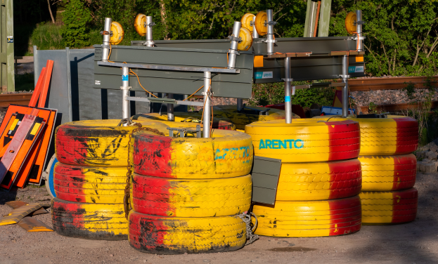 Ein Haufen gelber und roter Reifen auf einem Schmutzfeld umgeben von verstreuten Metallgegenständen, Brettern und anderem Schutt, mit Bäumen, Pflanzen und einer Hütte im Hintergrund.