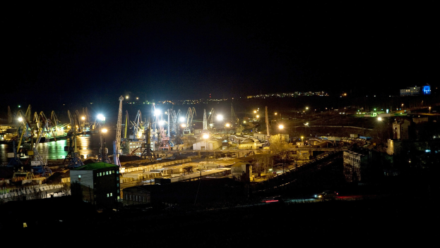 Eine nächtliche Stadtansicht mit beleuchteten Gebäuden, fahrenden Fahrzeugen auf einer Straße und einem Gewässer, vor einem dunklen Himmel.