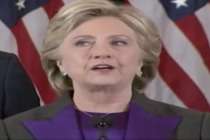 Hillary Clinton, in einem lila Kleid und schwarzer Blazer, spricht an einem Podium neben einem Mann, vor einer amerikanischen Flagge, mit entschlossener Miene.