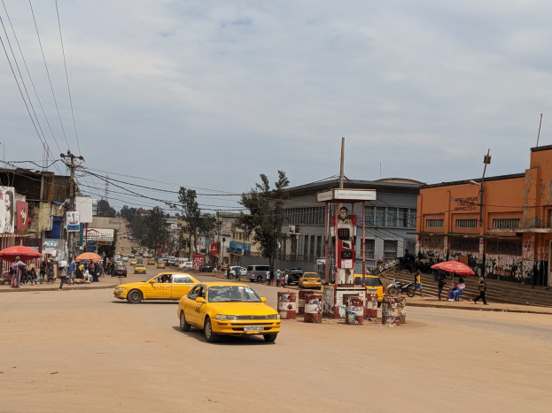 Ein gelbes Taxi fährt auf einer belebten Stadtstraße mit Gebäuden an den Seiten, während Fußgänger mit Schirmen auf dem Gehweg unter bewölktem Himmel gehen.