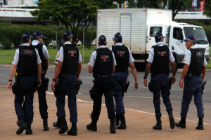 Eine Gruppe von Beamten in Uniform, einschließlich Mützen, Hemden, Schutzwesten, Uhren, Hosen und Schuhen, die auf einer Straße mit einem geparkten Lkw auf der rechten Seite neben einem Zaun gehen. Im Hintergrund sind Gebäude, Bäume, Pflanzen, Pfosten, Banner und eine Brücke im oberen rechten Eck zu sehen.