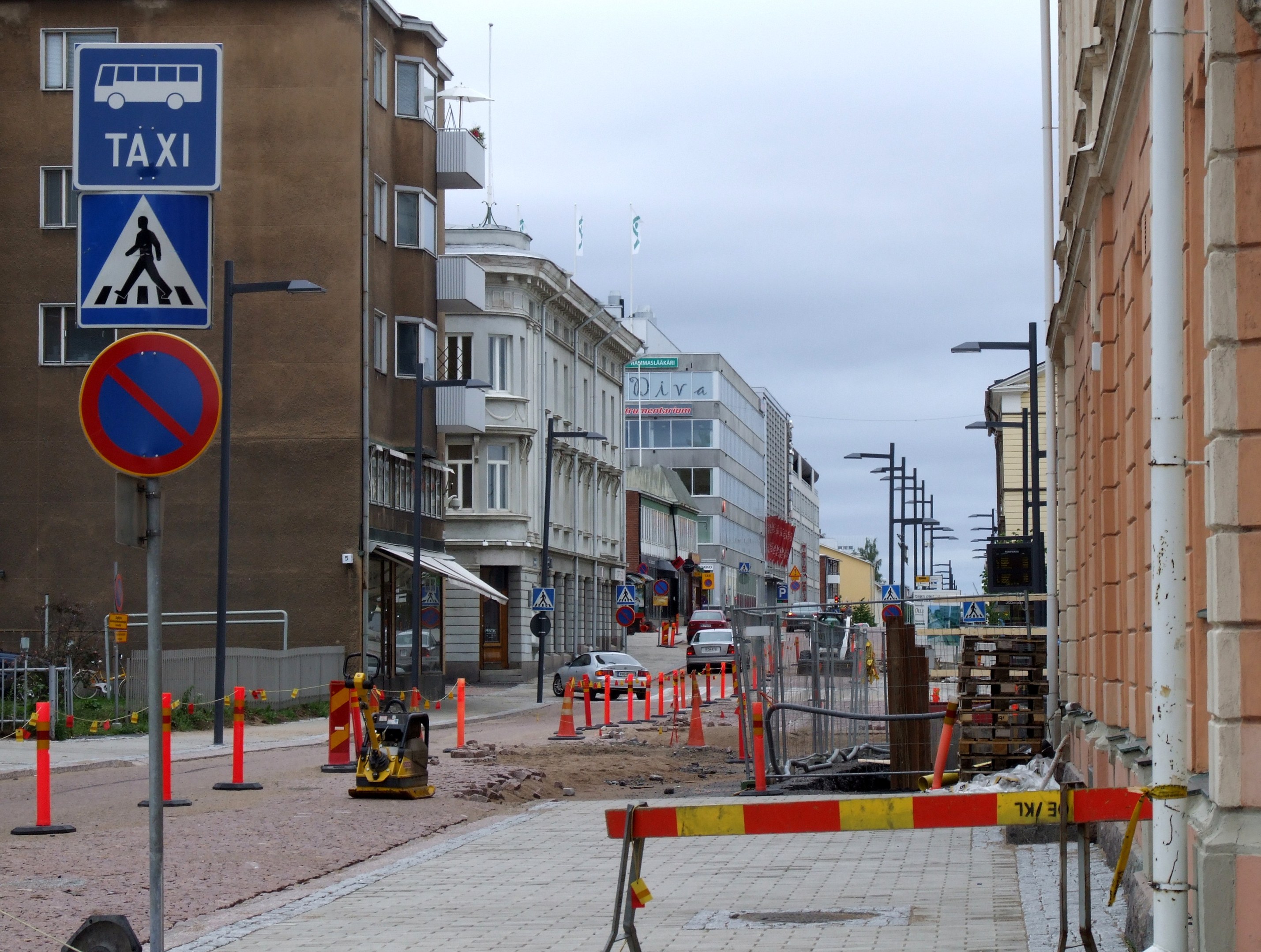 Stadtstraße mit Gebäuden, Straßenlaternen, Verkehrskegeln, Schildern, Fahrzeugen, Absperrpollern, Bäumen und einer Baustelle mit Verkehrszeichen unter einem bewölkten Himmel.