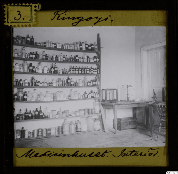 Schwarzes und weißes Foto einer alten Apotheke mit Regalen voller Flaschen, einem Tisch mit Gegenständen, einem Stuhl, einer Tür und einer Wand, mit Text unten.