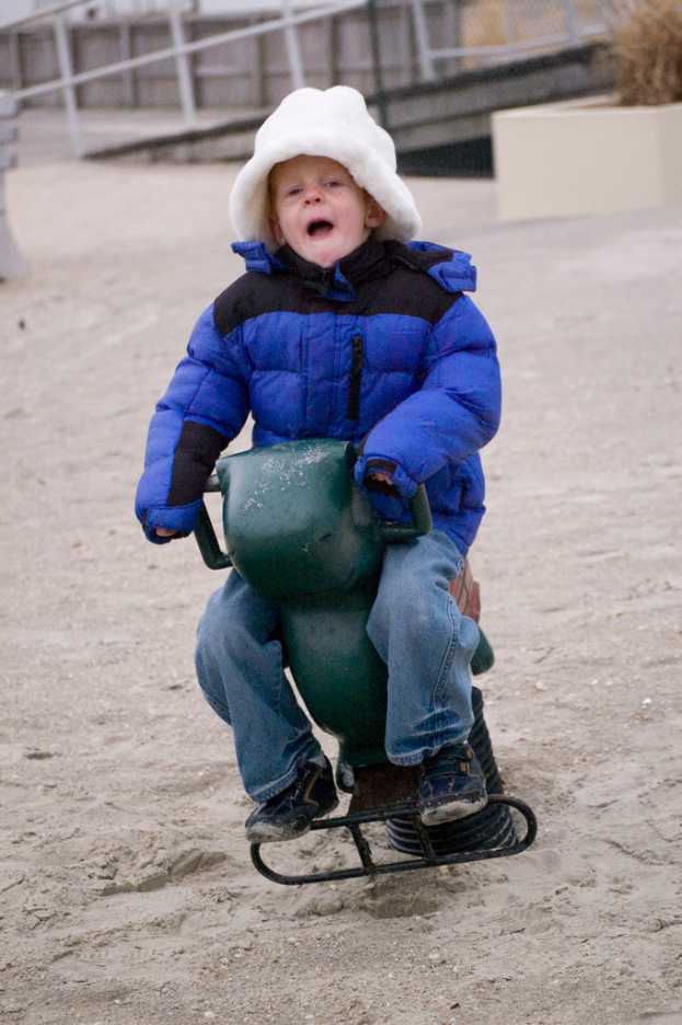 Ein kleiner Junge in einer blauen Daunenjacke sitzt auf einem Eisenfedersessel mit einem weißen Rohrgeländer im Hintergrund.