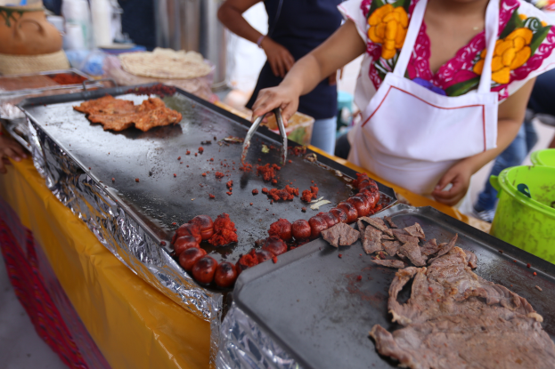 Eine Frau in einer Schürze kocht auf einem Grill an einem Markt, mit einem Tisch voller Lebensmittel, einem Eimer und anderen Gegenständen in der Nähe und einigen Menschen im Hintergrund.