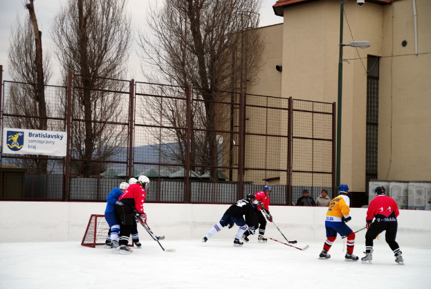 Menschen beim Eishockeyspielen auf einem Eisring mit Gebäuden, Bäumen, einer Straßenlaterne, einem Namensschild und Zäunen im Hintergrund unter einem klaren Himmel.