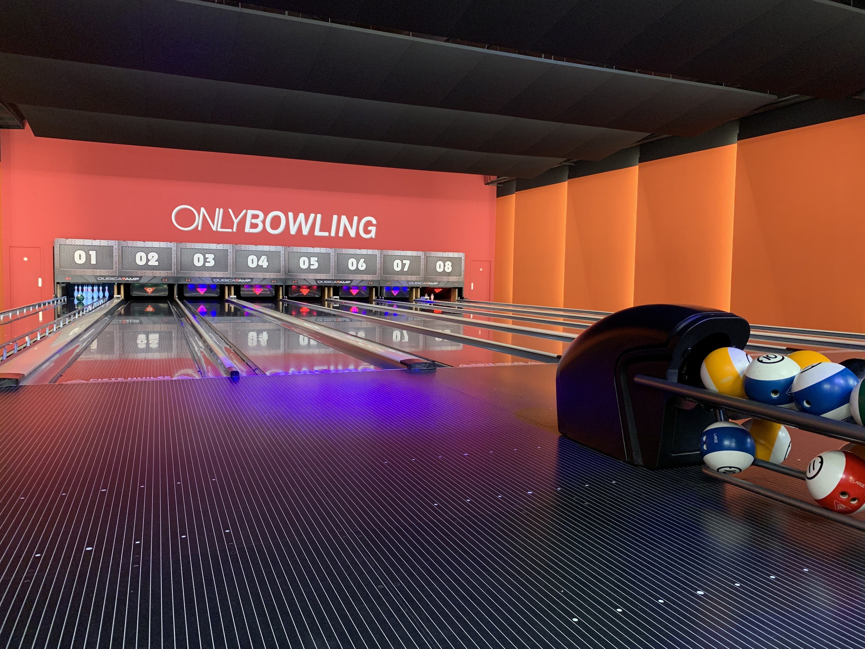 Eine Bowlinganlage mit Bowlingkugeln auf der rechten Seite, Kegel in der Mitte und einer Wand mit Text im Hintergrund, unter einer sichtbaren Decke.