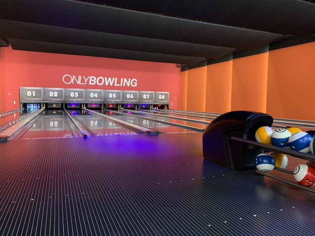 Eine Bowlinganlage mit Bowlingkugeln auf der rechten Seite, Kegel in der Mitte und einer Wand mit Text im Hintergrund, unter einer sichtbaren Decke.