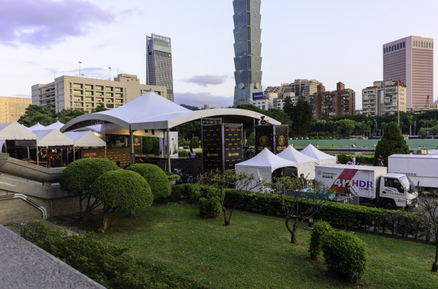 Eine Gruppe von Zelten in einem Park mit Grünflächen, Menschen, Fahrzeugen, Gebäuden und einem klaren Himmel, wahrscheinlich beim Hongkonger Filmfestival.