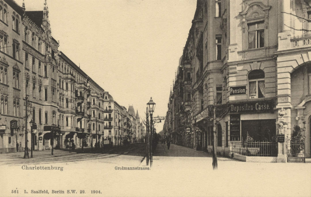 Schwarz-weiß-Foto einer Stadtstraße in Charlottenburg, Deutschland, mit Gebäuden, Straßenlaternen, Schildern, Kraftfahrzeugen, Fußgängern und Gittern unter einem klaren Himmel.