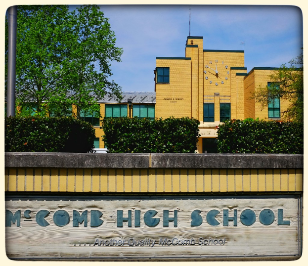 Ein Plakat mit der Aufschrift "MCCOMB HIGH SCHOOL" auf einer Wand, mit Pflanzen, Bäumen, einem Pfahl, einem Gebäude und einem bewölkten Himmel im Hintergrund.