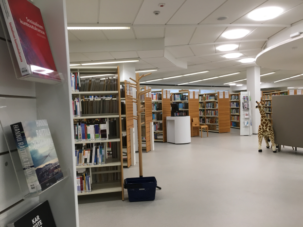 Ein geräumiger Bibliotheksinnenraum mit zahlreichen Bücherregalen, die mit Büchern gefüllt sind, ein Korb auf dem Boden, ein Giraffen-Spielzeug, Stühle, Tische, Säulen und Deckenleuchten.