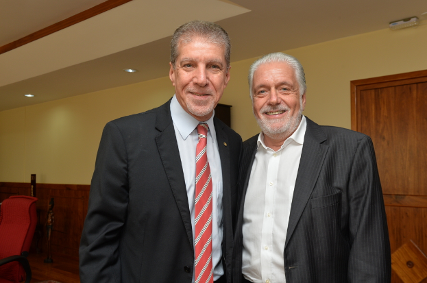 Zwei Personen in Anz├╝gen stehen und l├Ącheln vor einer gelben Wand mit brauner T├╝r, ein roter Stuhl ist links im Bild zu sehen.