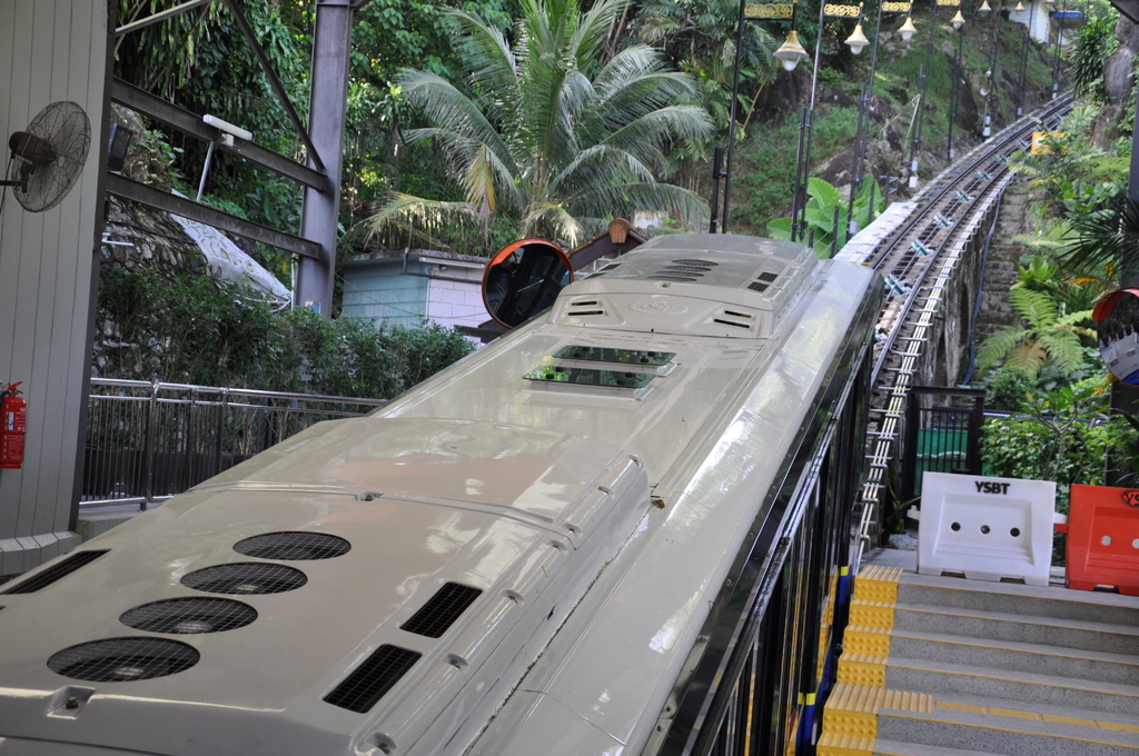 Ein Zug auf Eisenbahnschienen mit Gebäuden, Masten, einem Ventilator, einem Feuerlöscher und einem Geländer auf der linken Seite, verschiedene Gegenstände und eine Treppe auf der rechten Seite und Bäume im Hintergrund.