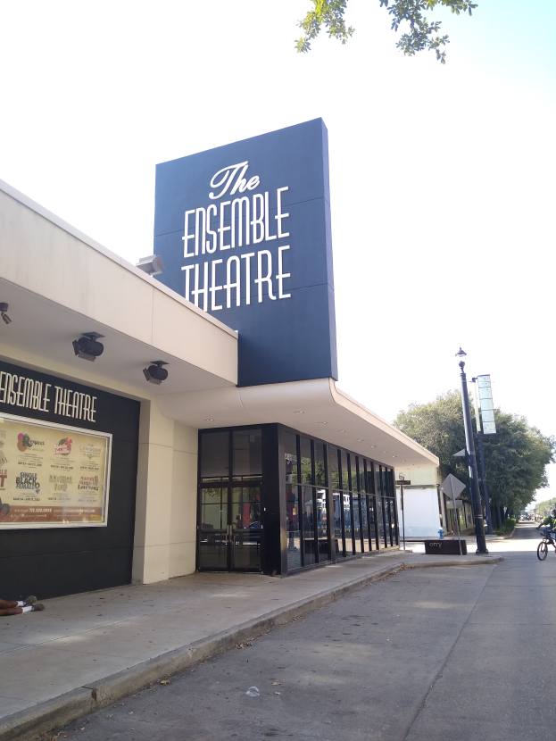 The Ensemble Theatre in San Francisco, a modern building with glass walls and doors, featuring a signboard, a person sitting on the footpath, a bicycle on the road, a street pole, trees, and a cloudy sky.
