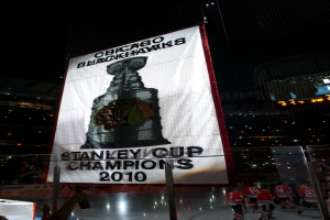 Eine nächtliche Stadion-Szene mit einem großen Banner in der Mitte, Zuschauern im Hintergrund, einem Glas auf dem Boden und einigen Spielern auf dem Feld, beleuchtet von Stadionlampen.