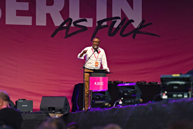 Ein Mann in weißem Hemd und braunen Hosen steht auf einer Bühne und hält ein Mikrofon in der Hand, mit einem pinken Banner im Hintergrund, das 'Berlin as Fuck' lautet und Lautsprecher auf der Bühne.