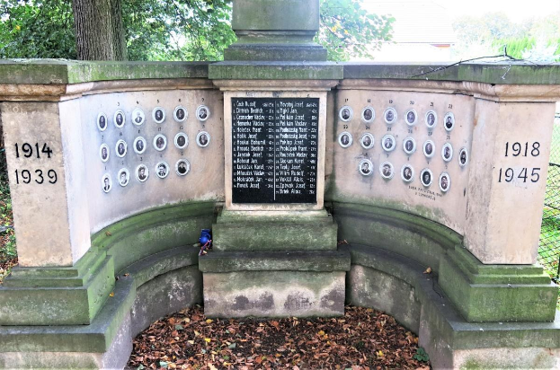 Ein Holocaust-Gedenkdenkmal in einem jüdischen Friedhof in Berlin, das eine Wand mit Text und Zahlen sowie umgebende Bäume und einen Zaun mit verstreuten trockenen Blättern zeigt.