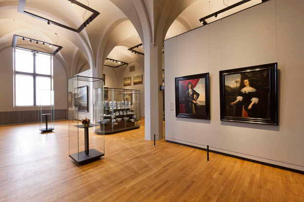 Ein geräumiger Galerie-Raum mit zahlreichen Gemälden, gerahmten Fotografien, Glasvitrinen mit Objekten, Ständern und Deckenleuchten, mit einem Wasserzeichen der National Gallery of Art.