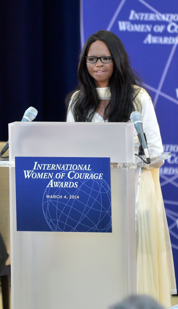 Frau In Weißen Kleid Und Brille, Die Auf Einem Podium Mit Zwei Mikrofonen Und Einer Tafel Steht, Vor Einer Menge, Mit Einem Blauen Banner Im Hintergrund, Das 'Internationale Frauen Der Courage Awards 2014' Bezeichnet Ist.
