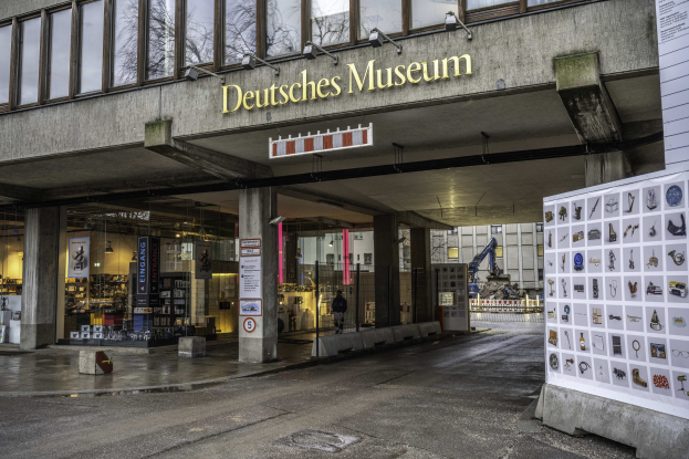 Außenansicht des Deutschen Museums in Berlin mit einem großen Gebäude mit Glasfenstern, Säulen und einer Namensplakette sowie einer Informationsplakette, umgeben von Bäumen, anderen Gebäuden und dem Himmel.