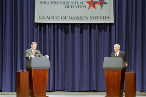 Zwei Männer in Blazern und Krawatten stehen an Podien mit Mikrofonen und Gläsern vor einem blauen Vorhang und einem Banner mit Text und Sternen, diskutieren die Debatten von 1984.