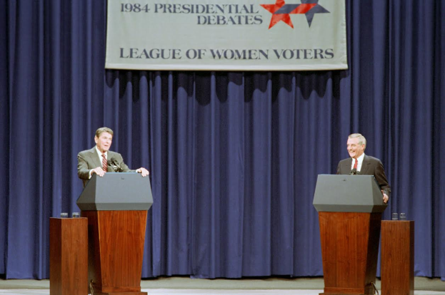 Zwei Männer in Blazern und Krawatten stehen an Podien mit Mikrofonen und Gläsern vor einem blauen Vorhang und einem Banner mit Text und Sternen, diskutieren die Debatten von 1984.