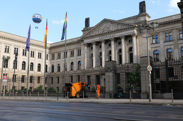 Großes modernes Gebäude mit vielen Fenstern, als Bundestag in Berlin, Deutschland identifiziert, umgeben von Straßeninfrastruktur und Fußgängern, mit Flaggen, Bäumen und einem bewölkten Himmel im Hintergrund.