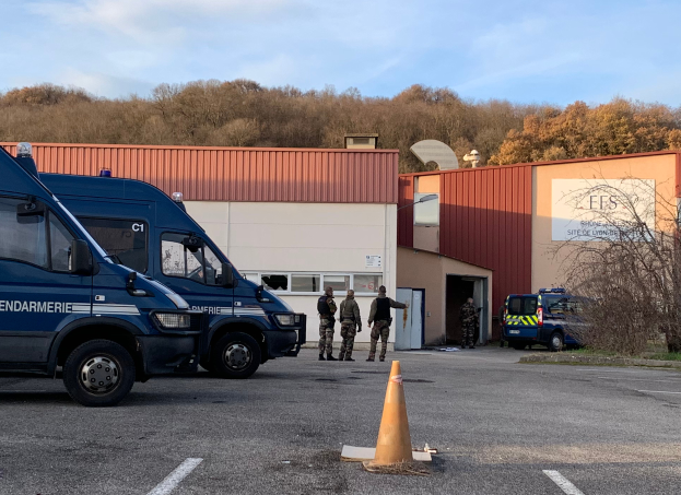Eine Gruppe von Polizeibeamten steht vor einem Gebäude mit Fenstern und Türen, umgeben von Fahrzeugen und einer Verkehrsinsel, mit Bäumen und einem bewölkten Himmel im Hintergrund.
