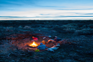 Ein Lagerfeuer mit Menschen, die darauf sitzen und liegen, mit Mötzen tragend, mit Tasche und anderen Gegenständen auf dem Boden, umgeben von Bäumen und Himmel.
