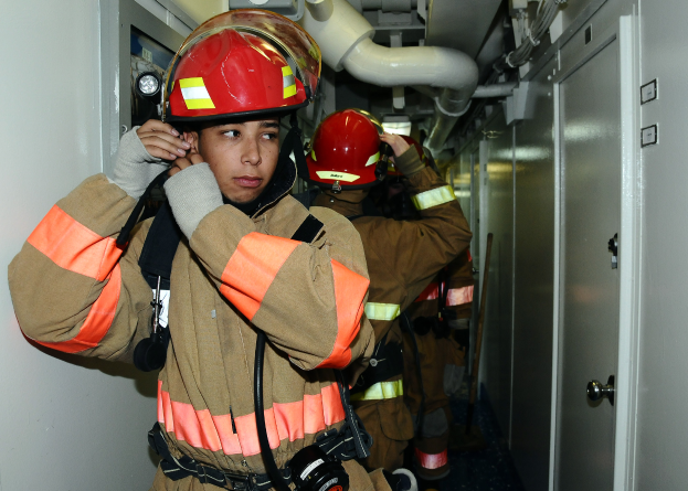 Eine Gruppe von Feuerwehrleuten in Uniform, die zusammen in einem Raum mit einer Tür auf der rechten Seite und einer Wand auf der linken Seite stehen, mit Rohren und anderen Gegenständen im Hintergrund.