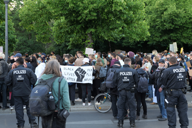 Eine große Gruppe von Menschen nimmt an einer Black-Lives-Matter-Demonstration in Berlin teil, einige halten Schilder und andere tragen Mützen und Taschen, mit einem Fahrrad im Vordergrund und Bäumen und einem Pfahl im Hintergrund.