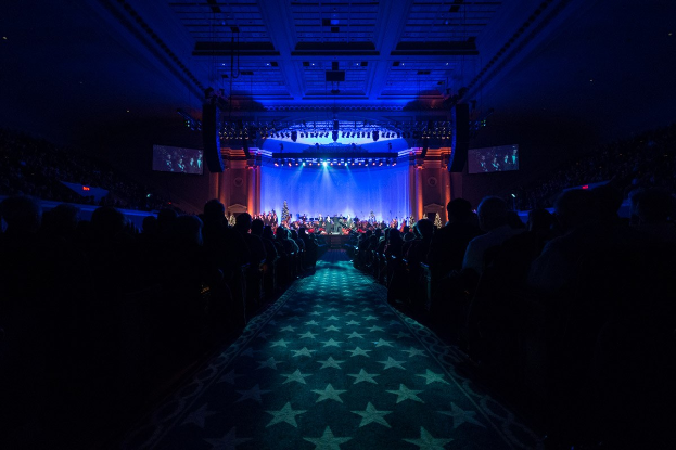 Ein großer Saal mit Stühlen, auf denen Menschen sitzen, die einer Bühne mit Musikern zugewandt sind, unter einer Decke mit Lampen und Fernsehgeräten, was auf eine Konzertsäleinrichtung hinweist.