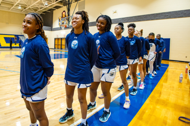 Gruppe junger Frauen in blauen T-Shirts, die auf einem Basketballfeld lächeln, mit Flaschen auf dem Boden, Zuschauern im Hintergrund und einem Basketballkorb.