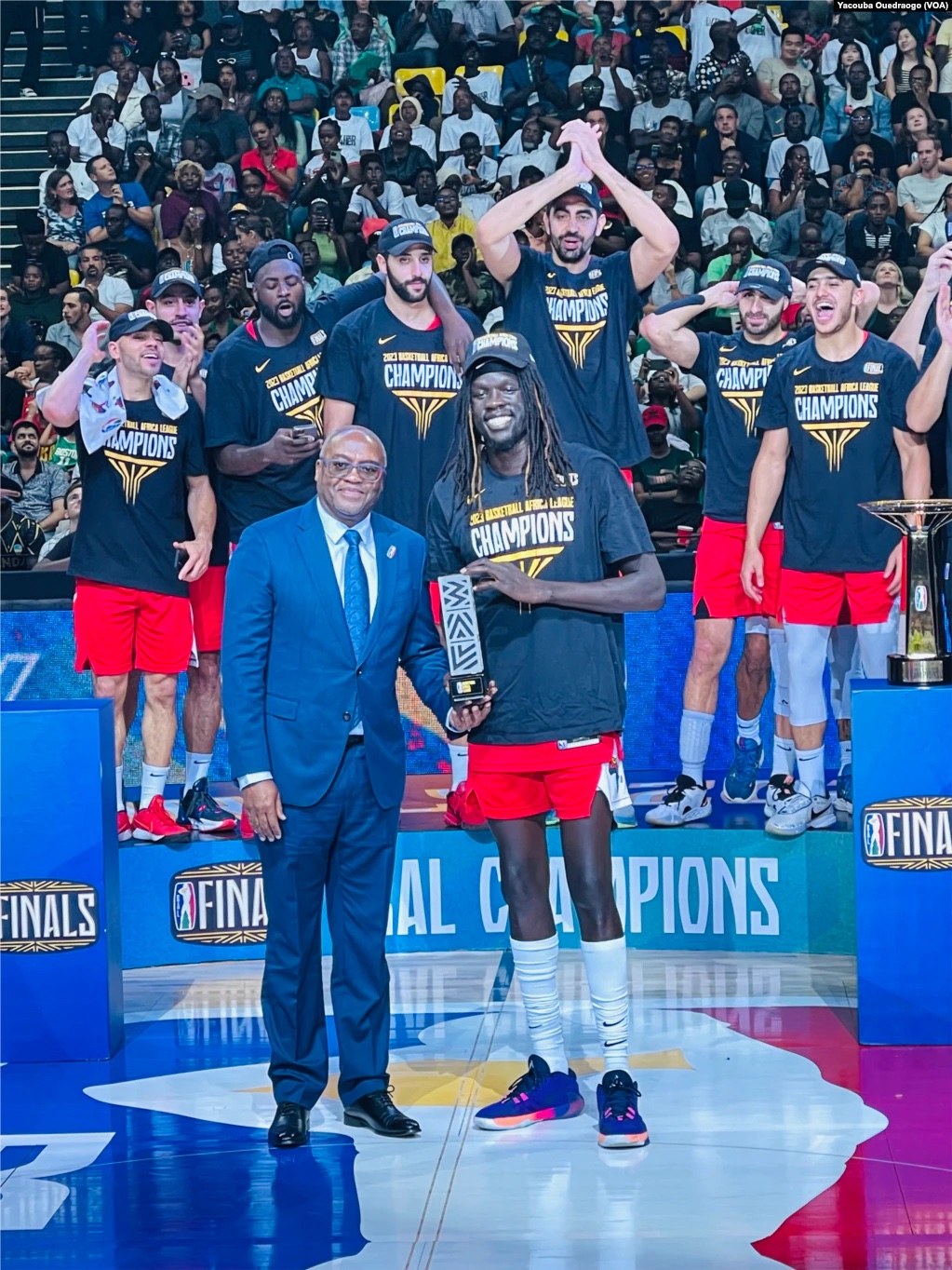 Männer mit Auszeichnungen auf einem Basketballfeld mit Zuschauern im Hintergrund und Auszeichnungen auf Podesten, die einen Sieg feiern.