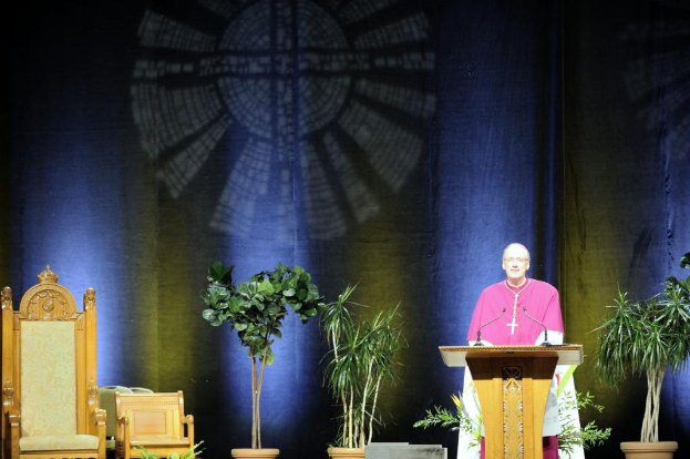 Ein Mann in einem rosa-weißem Kleid steht neben einem Rednerpult in einem Auditorium, mit Pflanzen und zwei Stühlen daneben und einer bunten Wand im Hintergrund.