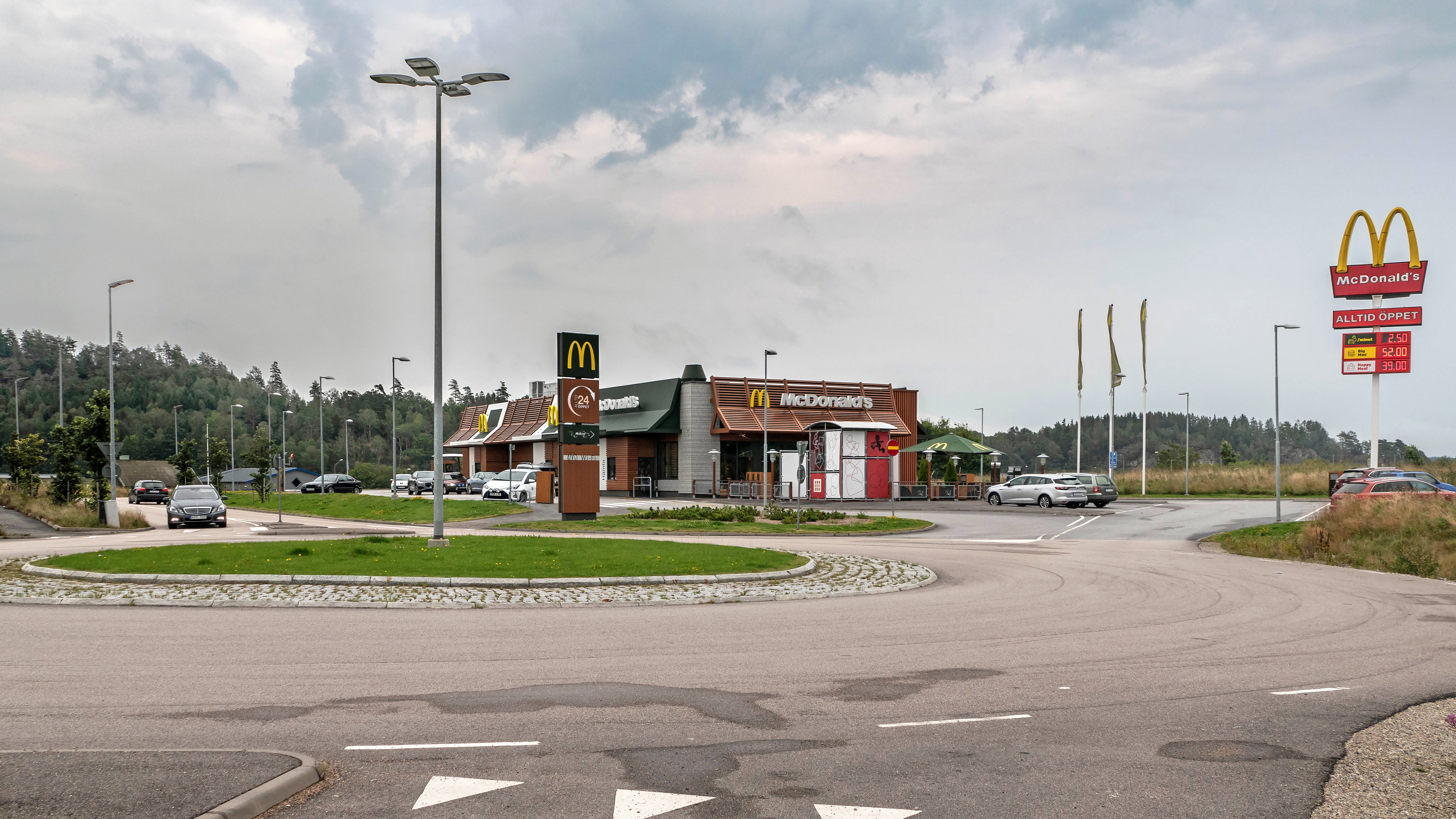 Ein McDonald's-Restaurant an einer Straßenecke, umgeben von Gebäuden, Straßenlaternen, Fahrzeugen, Infotafeln, Büschen, Bäumen und einem bewölkten Himmel.
