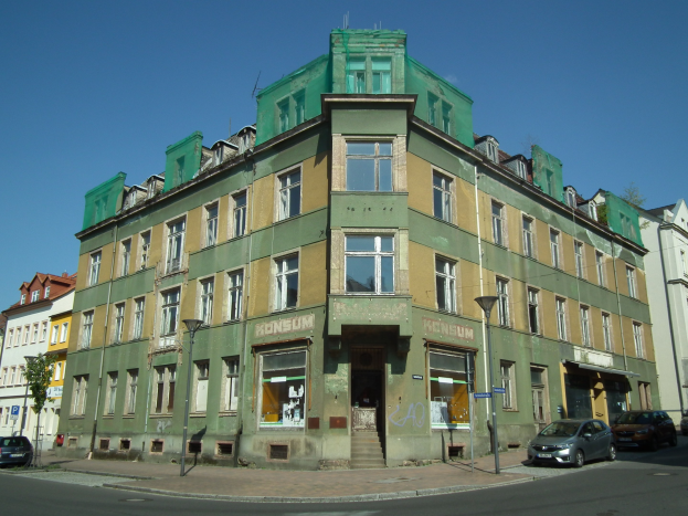 Altes Gebäude mit einer grünen Fassade an einer Straßenecke, umgeben von Straßenmasten, Straßenlaternen, Kraftfahrzeugen, Bäumen und einem klaren blauen Himmel.
