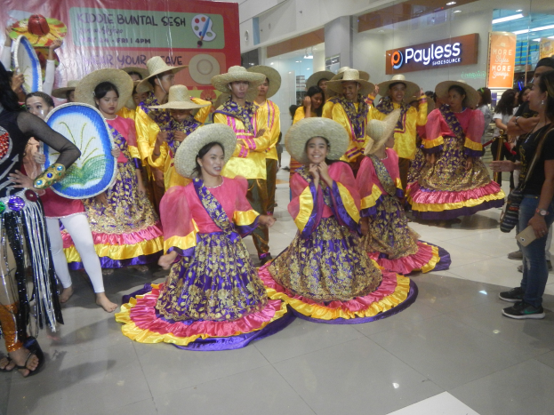 Eine Gruppe von Menschen in bunten Küsten und Hüten, die in einem Einkaufszentrum tanzen, mit anderen, die Musikinstrumente halten und ein Banner im Hintergrund zu sehen ist.