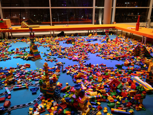 Ein großer Raum mit bunten Lego Bauklötzen, die auf dem Boden verstreut sind, umgeben von Tischen und Säulen, mit Glasfenstern im Hintergrund, die Menschen und Lichter zeigen.