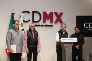 Vier Männer stehen nebeneinander vor einem Podium mit einem Mikrofon und einer Tafel mit der Aufschrift "CDMX, Mexikos erste digitale Währungsbörse"; zwei halten Akten, zwei tragen Brillen, mit Flaggen und einem Bildschirm im Hintergrund.